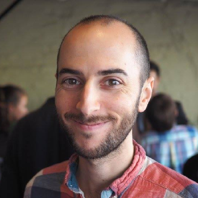 Headshot of a smiling, bearded white man in his 30s, wearing a colourful plaid shirt